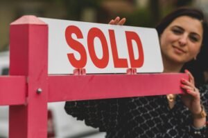 About Woman smiling behind a 'Sold' sign, symbolizing successful home purchase.